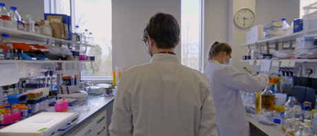 Man wearing lab coat walking between two lab benches. 