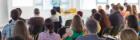 Heads of an an audience seen from behind as they listen to a speaker standing in front of them. Source: Shutterstock