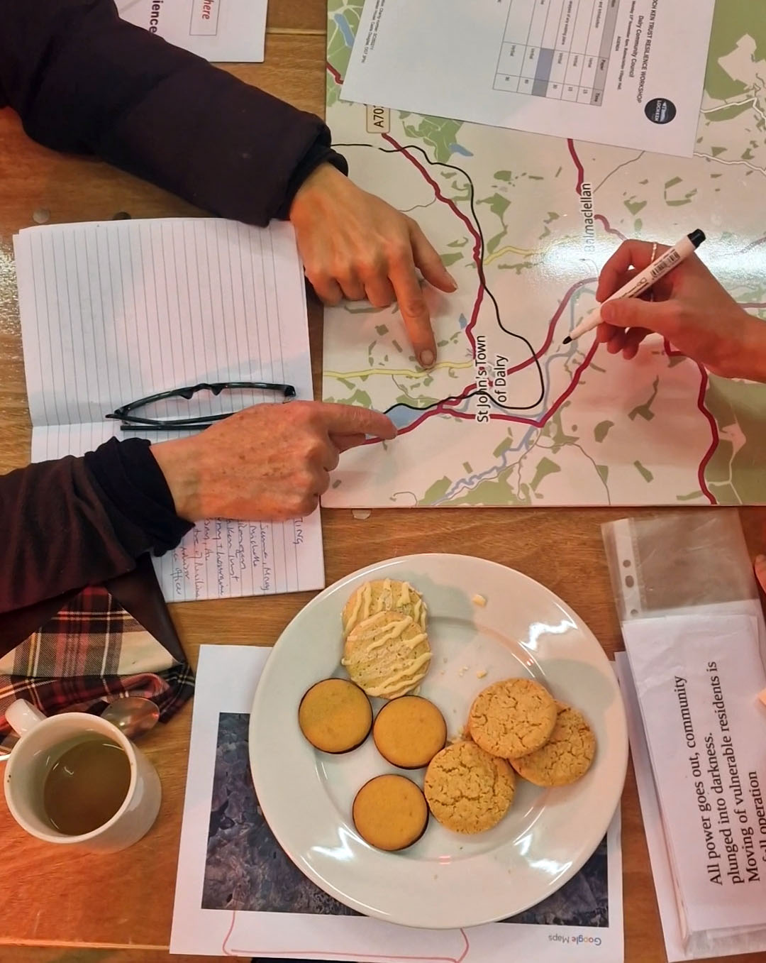 Image shoes hands pointing at a map and someone writing points on the map with a pen. Sat on the table are also cups of tea and biscuits. 