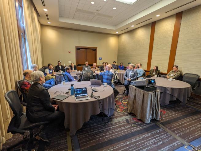 A room full of people sat at 3 round tables in a meeting room. 