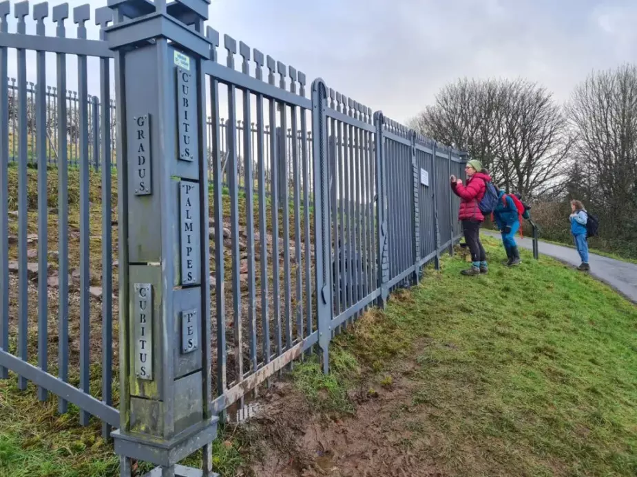 Alison Phipps, Bella Hoogeveen and Jennifer McArthur looking into gated enclosure containing small excavated area of the Antonine Wall