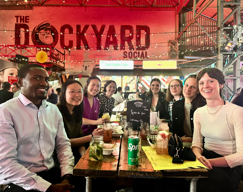 A group shot of the Clinical Viral Epidemiology Group sat at a table in a bar all facing towards camera