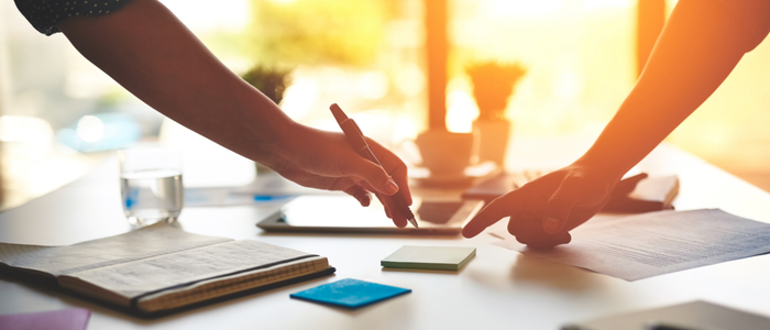 Two hands holding pens above a table with notebooks and post its.