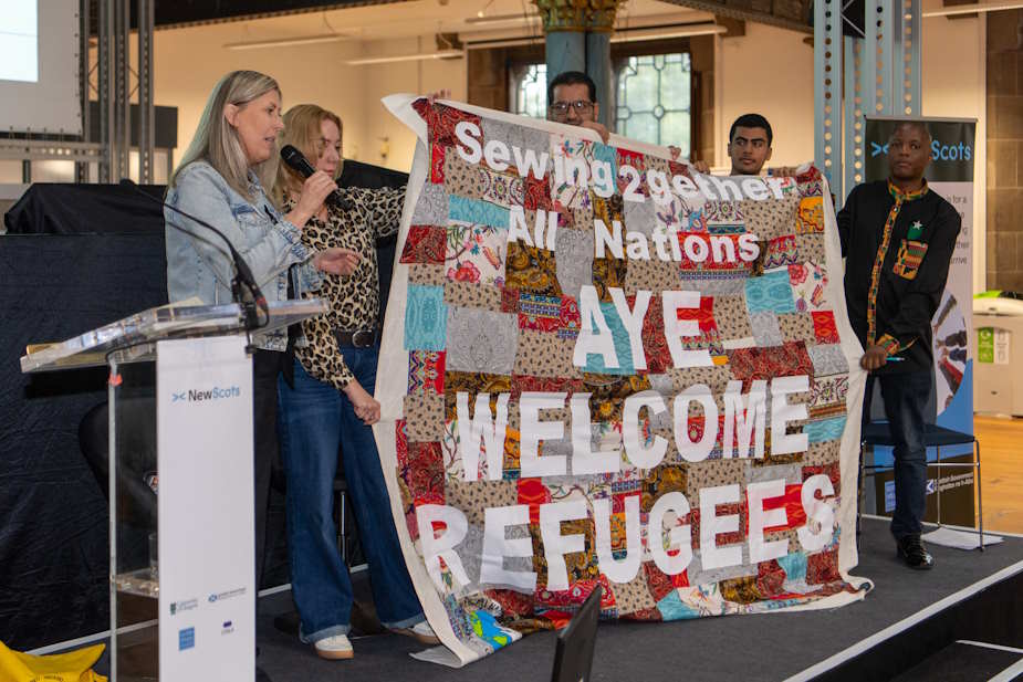 Group of people holding patchwork blanket reading Sewing 2gether All Nations AYE WELCOME REFUGEES