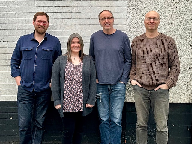 Three men and a woman, all very good looking standing together against a white wall