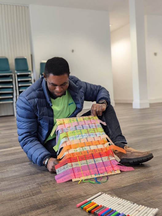 Person sitting on the ground holding up board with woven strips of coloured paper with words written on them