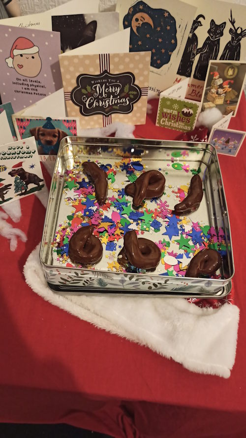 Photo of a silver tin containing multicoloured confetti and plastic fake excrement. The tin is sitting on top of a Santa hat on a table and surrounded by Christmas cards.