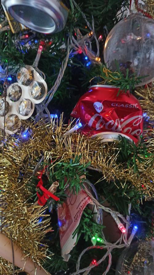 Close up photo of the branches of a Christmas tree decorated with lights, tinsel and assorted rubbish including a crushed Coca-Cola can.