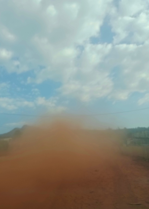 Photo of a cloud of red dust rising from a road with a blue sky and clouds above.