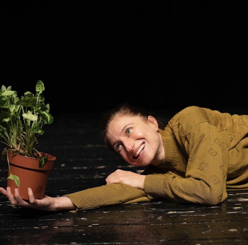 Actress Nezaket Erden lying on her side on the ground looking towards a potted plant held in her outstretched hand.