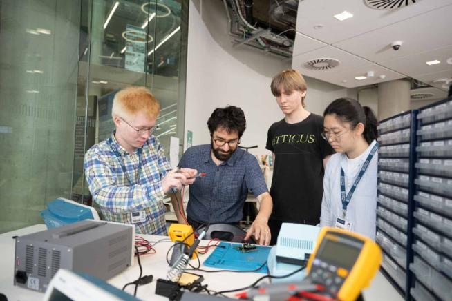 A group at a bench building some electronics with a soldering iron and test equipment around them
