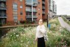 Woman standing in front of a flower garden and a block of flats, wearing a white jumper, black skirt and tote bag