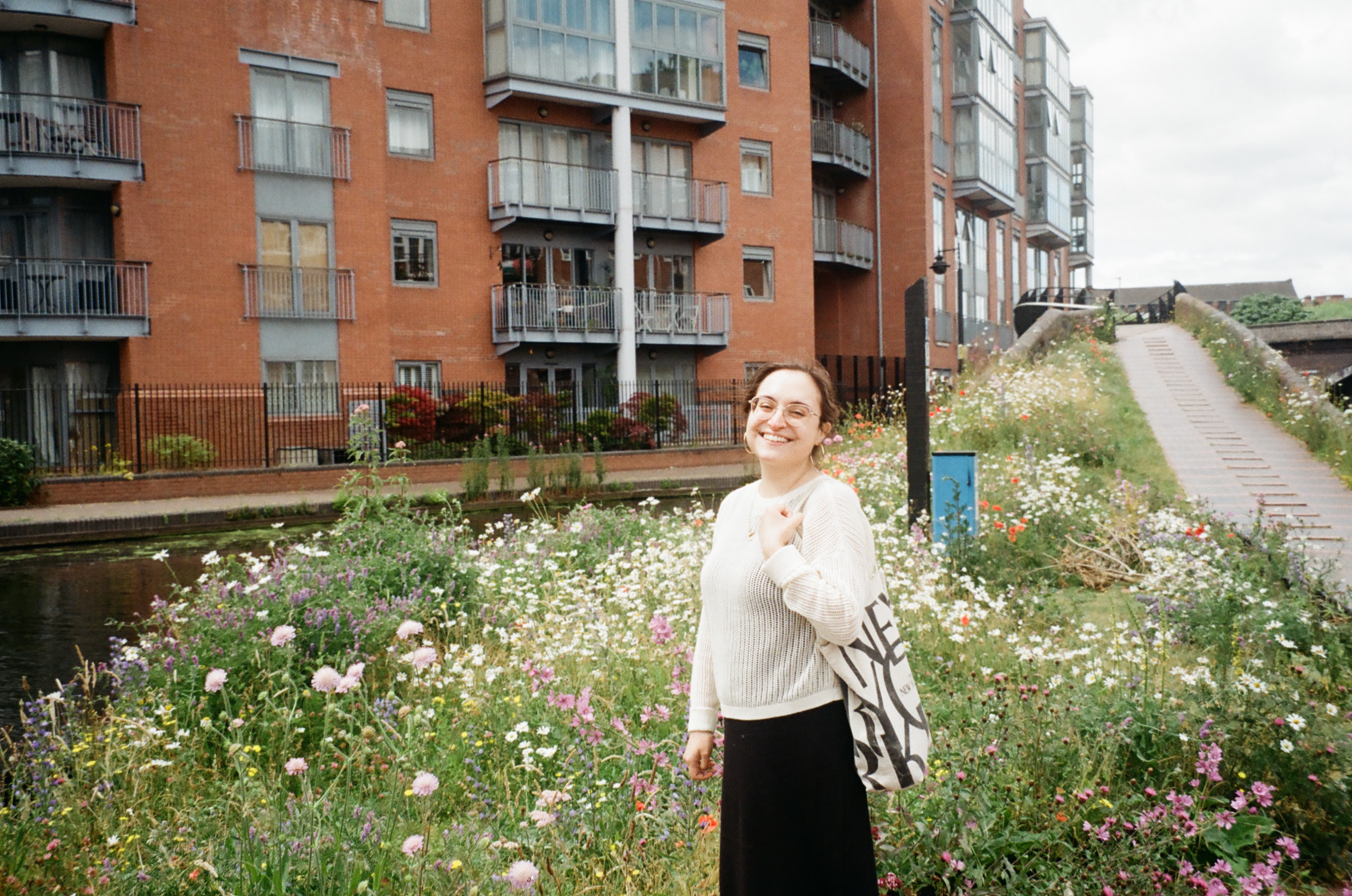 Woman standing in front of a flower garden and a block of flats, wearing a white jumper, black skirt and tote bag