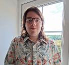 A man with shoulder-length hair, glasses, and a floral shirt, standing in front of a window