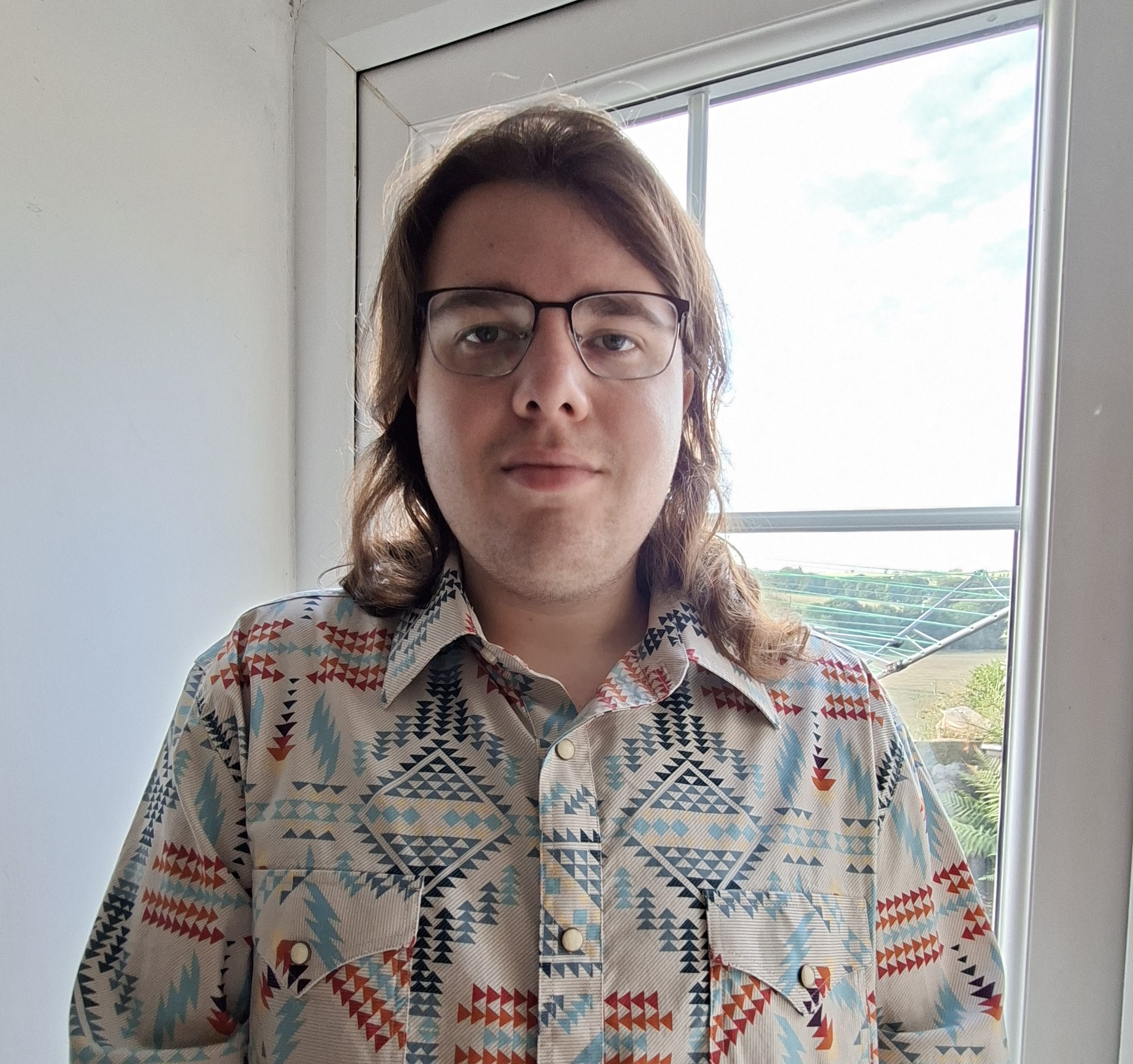 A man with shoulder-length hair, glasses, and a floral shirt, standing in front of a window