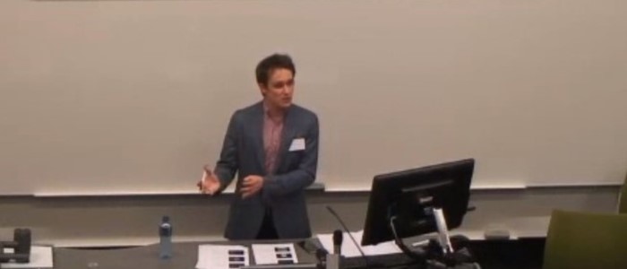 photo of a white man in a dark suit and pink shirt gesticulating while speaking in front of a desk and computer