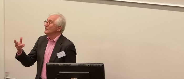 photo of an old white man with short white hair in a suit gesticulating while talking in front of a computer screen