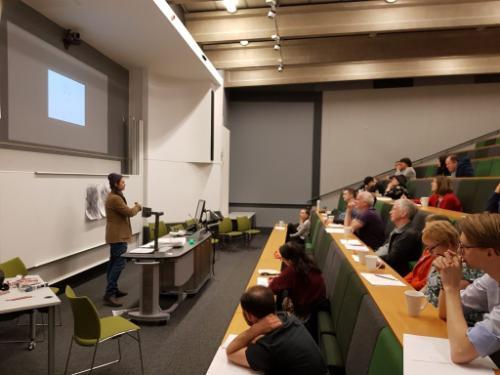 photo of a person with long hair and a beanie hat speaking in front of an audience in an auditorium, in front of a projector screen