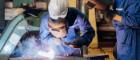 Two men in a factory; one is leaning down holding a tool that is creating a pure white heat and sparks above a workcounter and one is standing watching. They are both wearing boiler suits , gloves, hard hats and holding metal welding masks in front of their faces. Source: Phawat Topaisan | iStockPhoto  https://www.istockphoto.com/photo/group-of-technician-industrial-engineers-wearing-safety-unifor