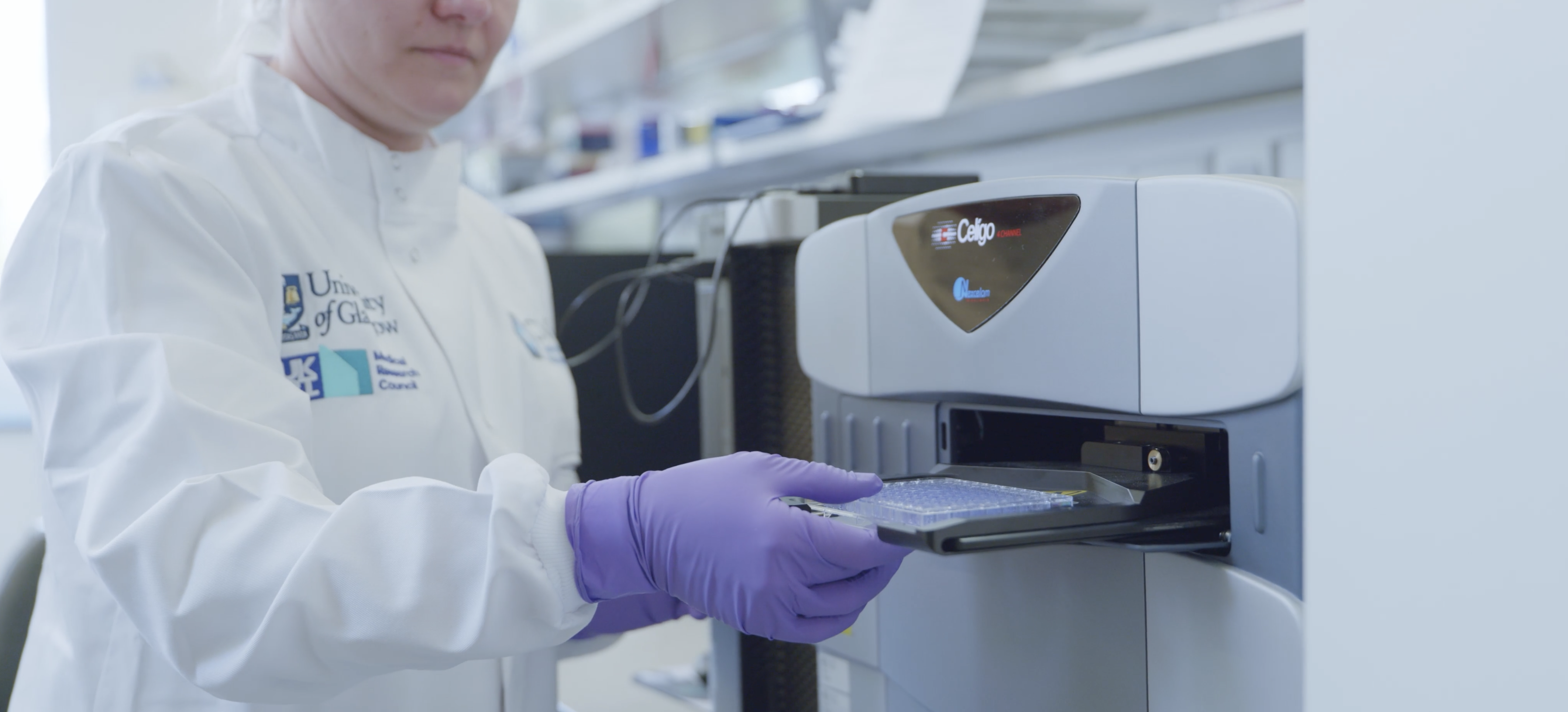 Person wearing lab coat and purple gloves inserting a tray into a machine in a laboratory.