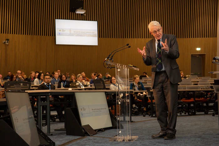 A speaker in front of an audience in a lecture hall, turning and gesticulating as he talks: Source: University of Glasgow