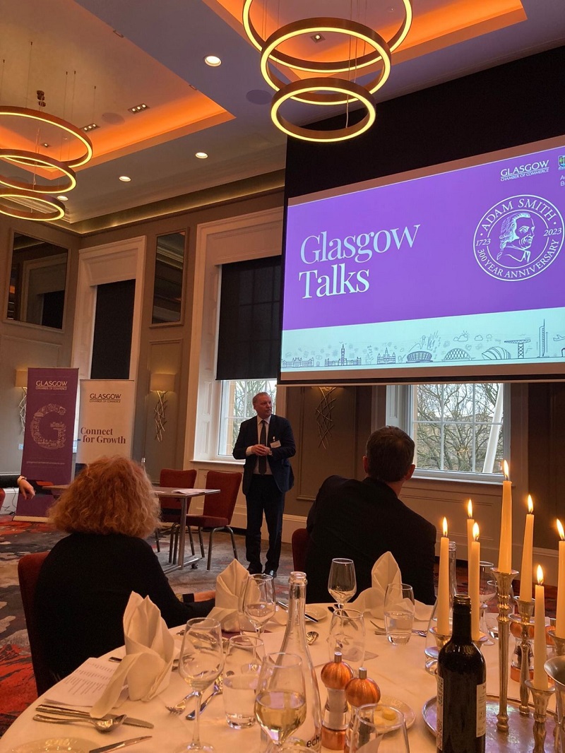 An event with groups of people at round dinner tables with white tablecloths and candles listening to a man giving a talk. Source: University of Glasgow