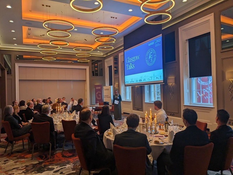 An event with groups of people at round dinner tables with white tablecloths and candles listening to a woman giving a talk. Source: University of Glasgow