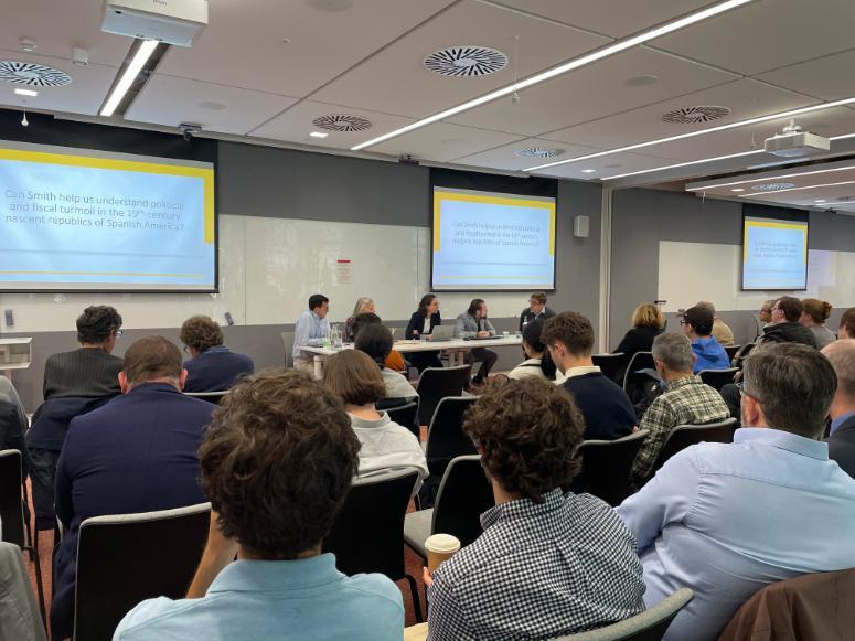 A crowd of people looking at the 5  lectures: Alex Trew, Deirdre McCloskey, Vladimir Maltse, Francesco Fiore and Luz Arias. With four whiteboards behind them introducing the main topic of discussion: Can Adam Smith help us understand the nature and causes of the wealth of nations in 2023. Source: Claire Martin, College of Social Sciences