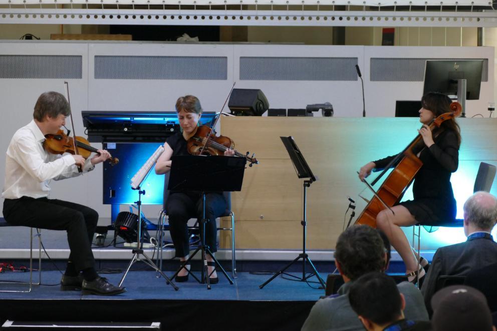 3 people play musical instruments. Two playing violins and one playing Cello with a blue light behind them. Source: ASBS