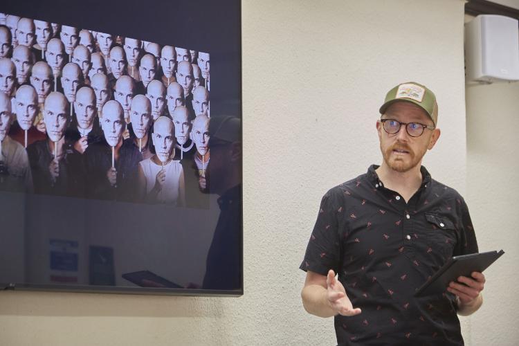 A presenter talking holding and ipad in one hand and has a screen behind him with people holding masks of the same person. Source: Charlotte Morris External Relations